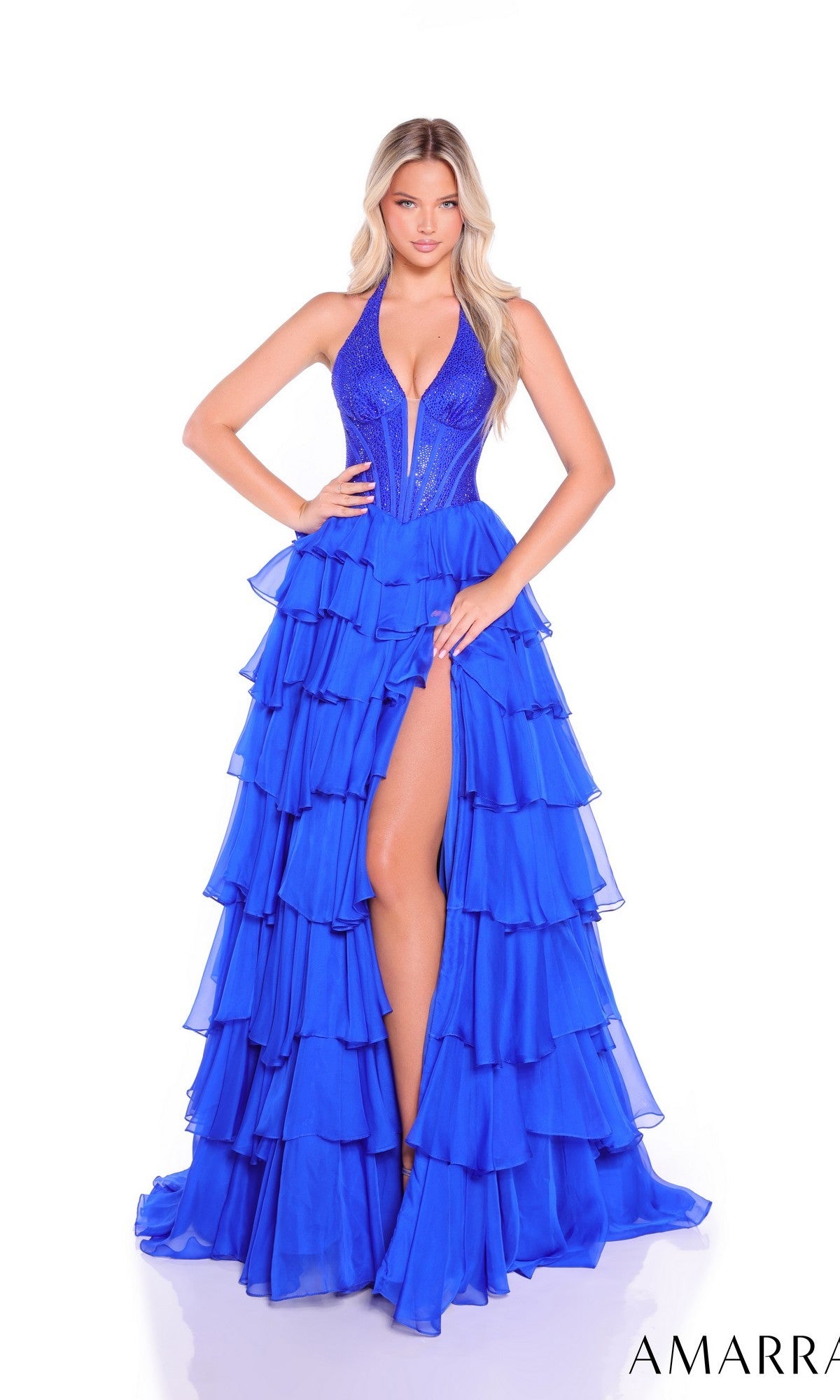 A woman wears the Amarra 89287 long prom dress, featuring a vibrant blue color, halter neckline, deep V front, high slit, and tiered chiffon A-line skirt, standing against a white background.